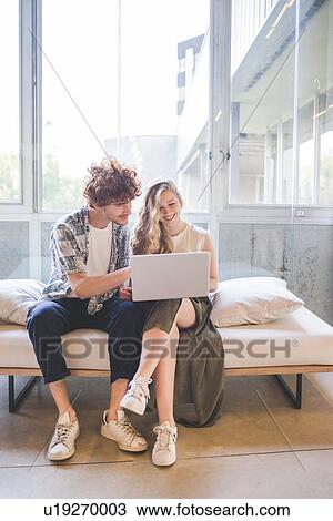 Stock Image - Co-workers working on laptop on sofa by window. Fotosearch