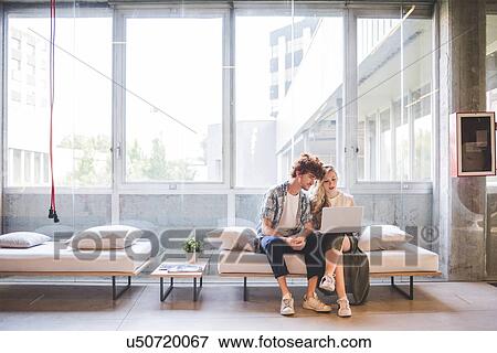 Stock Photo - Co-workers working on laptop on sofa by window. Fotosearch