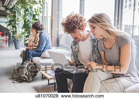 Stock Photo - Co-workers working on laptop on sofa by window. Fotosearch