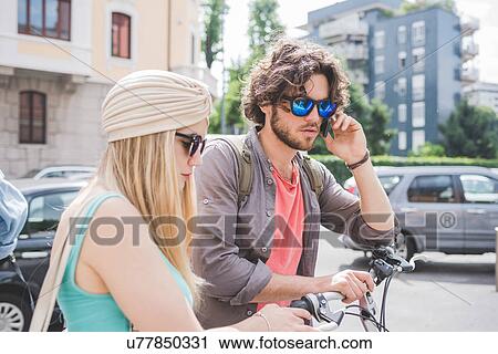 Stock Image - Couple using phone and walking along with push bikes. Fotosearch