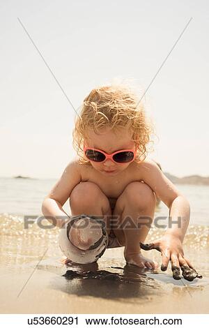Stock Image - Cute girl crouching on beach playing with plastic cup in sand, Alicante, Spain. Fotosearch