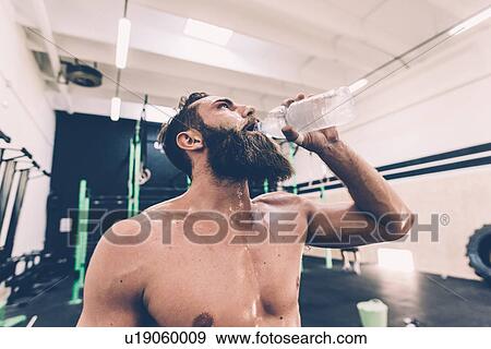 Stock Photo - Exhausted male cross trainer drinking water in gym. Fotosearch