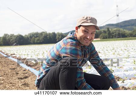 Stock Photo - Farmer in field tending to seedlings, looking at camera smiling. Fotosearch