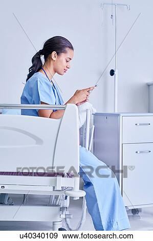 Stock Photo - Female medic sitting on hospital bed reading smartphone text messages. Fotosearch