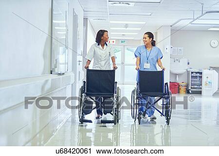 Stock Image - Female orderlies pushing wheelchairs along hospital corridor. Fotosearch