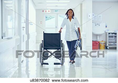 Stock Image - Female orderly pushing wheelchair along hospital corridor. Fotosearch