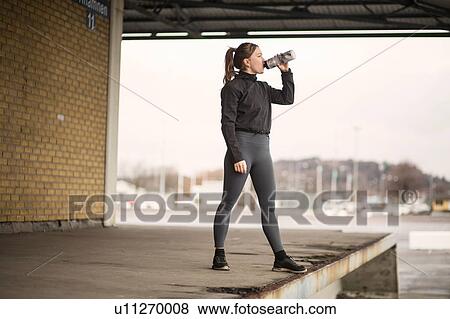 Stock Photo - Female runner drinking bottled water on warehouse platform. Fotosearch