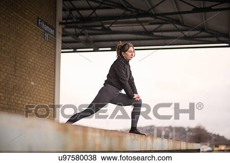 Stock Photo - Female runner leaning forward stretching on warehouse platform. Fotosearch