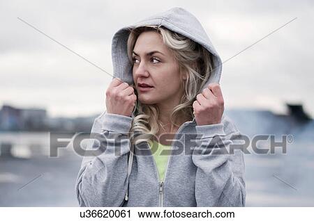 Stock Image - Female runner putting up hoody on rainy dockside. Fotosearch
