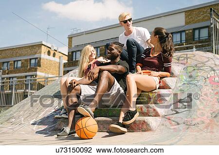 Stock Image - Four male and female basketball friends sitting chatting in city skatepark. Fotosearch