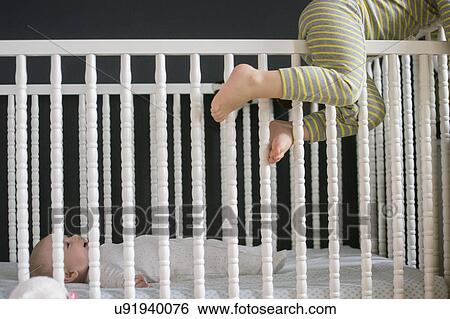 Stock Photograph - Girl (2-3) climbing on crib and brother (6-11 months) lying inside. Fotosearch