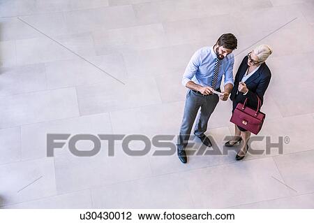 Stock Image - High angle view of two businessman and woman reading smartphone text in office atrium. Fotosearch