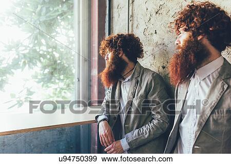 Stock Photo - Identical male hipster twins looking out of office window. Fotosearch