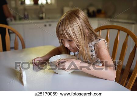 Stock Photo - Little girl eating breakfast while watching electronic device. Fotosearch