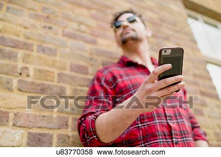 Low angle view of young man leaning against brick wall holding smartphone View Large Photo Image Stock Photo - Low angle view of young man leaning against brick wall holding smartphone. Fotosearch