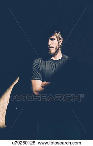 Stock Photo - Low key portrait of young man with folded arms staring from shadows. Fotosearch
