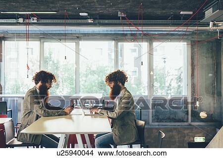 Picture - Male adult hipster twins sitting face to face at desk in office. Fotosearch