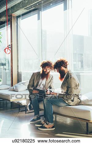 Stock Photography - Male adult hipster twins sitting whilst working on laptop in office. Fotosearch