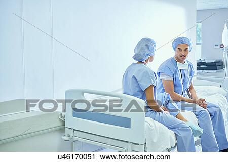 Stock Image - Male and female nurse sitting on bed chatting in hospital ward. Fotosearch