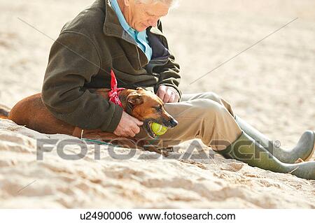 Stock Photograph - Man and dog sitting on beach, Constantine Bay, Cornwall, UK. Fotosearch