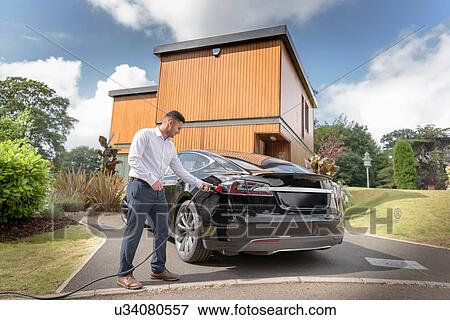 Stock Photo - Man charging electric car outside modern house. Fotosearch