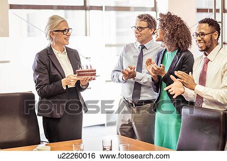 Stock Photograph - Mature woman presenting cake with candles to business team in boardroom. Fotosearch