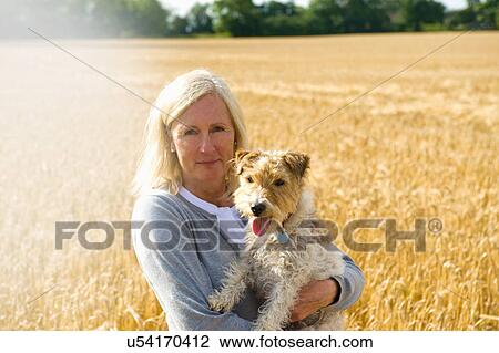 Stock Image - Mature woman standing in field, holding pet dog. Fotosearch