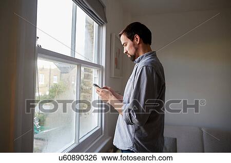 Stock Photograph - Mid adult man at living room window reading smartphone texts. Fotosearch
