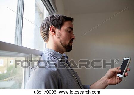 Stock Photograph - Mid adult man at window reading smartphone texts. Fotosearch