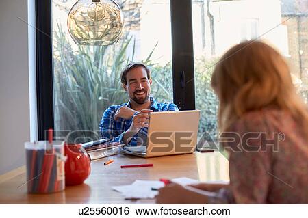 Stock Photograph - Mid adult man using laptop at dining table whilst daughter colouring. Fotosearch