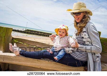Picture - Mid adult woman and baby daughter sitting on beach groyne, Ferring Beach, West Sussex, UK. Fotosearch