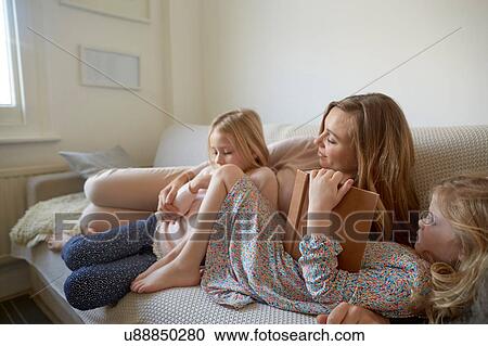 Stock Image - Mid adult woman and daughters reading storybook on sofa. Fotosearch