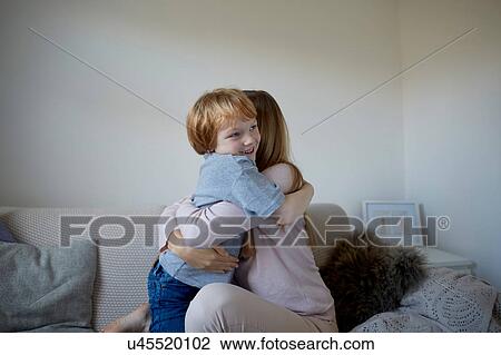 Stock Image - Mid adult woman and son hugging on sofa. Fotosearch