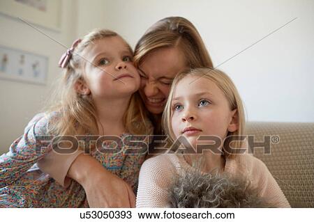 Stock Image - Mid adult woman hugging daughters in living room. Fotosearch