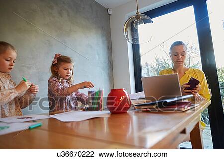 Mid adult woman reading smartphone texts at dining table whilst daughters colouring View Large Photo Image Stock Image - Mid adult woman reading smartphone texts at dining table whilst daughters colouring. Fotosearch