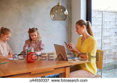 Stock Photo - Mid adult woman reading texts at dining table whilst daughters colouring. Fotosearch