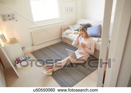 Stock Image - Mid adult woman sitting on living room floor using laptop. Fotosearch