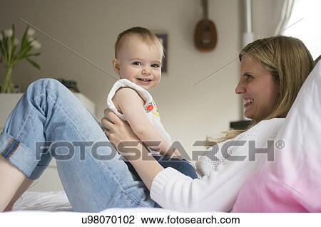 Mother and daughter (18-23 months) sitting on bed and smiling View Large Photo Image Stock Image - Mother and daughter (18-23 months) sitting on bed and smiling. Fotosearch