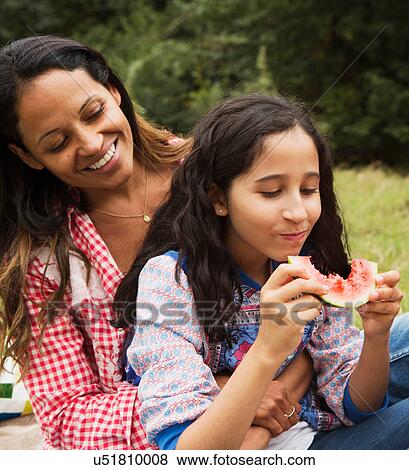 Stock Photo - Mother and daughter sitting outdoors, daughter eating watermelon. Fotosearch