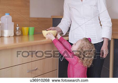 Stock Photo - Mother helping daughter (4-5) take bowl from kitchen counter. Fotosearch