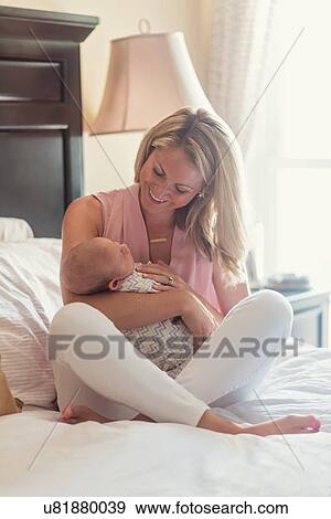 Stock Photo - Mother sitting on bed and holding baby son (0-1 months). Fotosearch