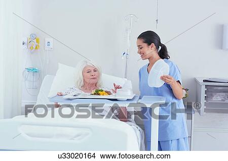 Picture - Nurse serving lunch to senior female patient in hospital bed. Fotosearch