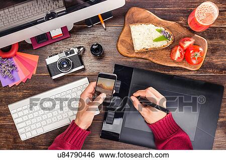 Stock Photograph - Overhead view of male hands editing photographs from smartphone on graphic design tablet using digital pen. Fotosearch