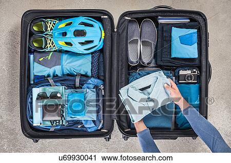 Stock Image - Overhead view of man's hands packing suitcase with walking boots, bike helmet, backpack, retro camera and blue shirt. Fotosearch
