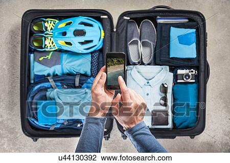Stock Image - Overhead view of man's hands using smartphone touchscreen above packed suitcase with blue bike helmet, backpack, retro camera and shirt. Fotosearch