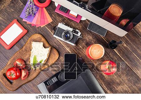 Stock Image - Overhead view of photo editing equipment; graphic tablet, retro camera, smartphone, desktop computer and lunch. Fotosearch
