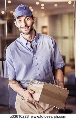 Stock Image - Portrait of delivery man carrying package in office. Fotosearch