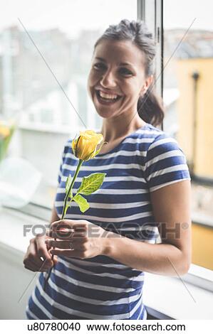 Stock Image - Portrait of happy young woman in apartment holding yellow rose. Fotosearch