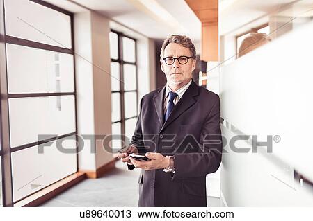 Stock Image - Portrait of senior businessman at office doorway using smartphone. Fotosearch