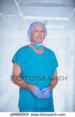 Stock Image - Portrait of senior male medic wearing scrubs in hospital corridor. Fotosearch
